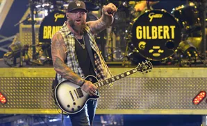 Guitarist with a beard, wearing a plaid shirt and a baseball cap, plays a white Gibson guitar on stage.