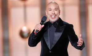Comedian Jo Koy in a black velvet tuxedo, holding a microphone and gesturing with his hand.