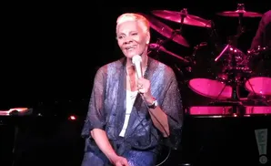 Dionne Warwick sings into a microphone on stage, wearing a shimmering robe over a white top.