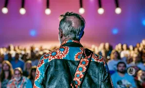 Back view of a man with a floral shirt and leather jacket, facing a crowd with purple and blue stage lights.