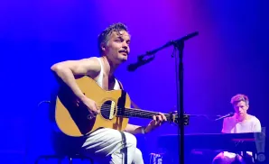 A man in a white tank top plays an acoustic guitar on a stage with a blue background. Another man plays a keyboard to his right.