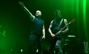 A bald singer with arm raised stands left of a guitarist playing a red electric guitar on a dimly lit stage.