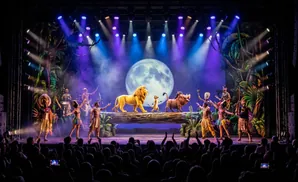 A lively stage performance of The Lion King with actors in animal costumes, puppetry, and live music.