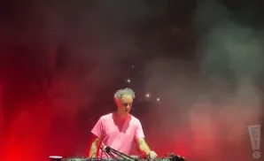 A man with curly gray hair in a pink t-shirt stands behind DJ equipment on a stage lit by red lights and smoke.