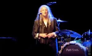 Patti Smith performs on stage with a drum kit behind her decorated with flowers.