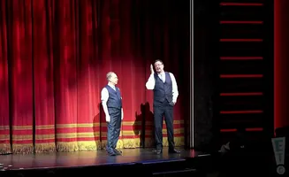 Two men in vests and ties stand on stage in front of red curtains.