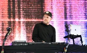 DJ wearing a black t-shirt and cap, standing behind DJ equipment with a laptop and speakers.