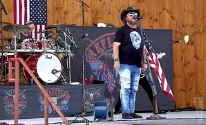 A country singer in a cowboy hat and distressed jeans performs with an American flag and skull design on his shirt.