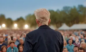 Back of an older man with white hair speaking to a large, blurred crowd outdoors.