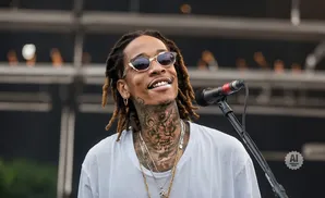 Rapper Wiz Khalifa smiles and performs into a microphone, wearing sunglasses and a white t-shirt with numerous necklaces.