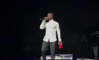 Comedian Kevin Hart on stage in a white shirt and dark pants, holding a microphone and a red cup.