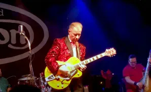 A man in a sparkly red jacket plays a yellow electric guitar on stage.