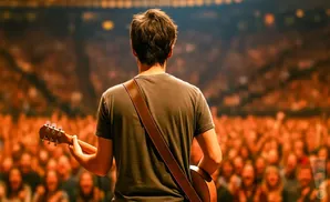 A guitarist on stage plays for a large, blurred audience, seen from behind.