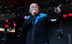 Musician with white hair wearing a black and blue shirt with a tambourine and microphone on stage.