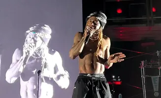 Rapper Wiz Khalifa performs on stage, shirtless, wearing a beanie and black shorts, with a large projection of himself behind him.