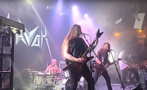 A band performs on stage with a large logo behind them.