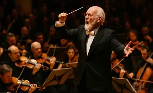 A conductor leads an orchestra, baton raised, with musicians playing violins in the background.