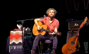 A man plays an acoustic guitar on stage, with a Marshall amp and another guitar beside him.