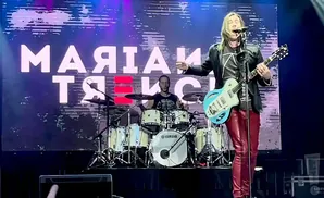 A man in a black jacket and red pants plays a blue guitar on stage with a drummer behind him.