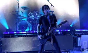 A guitarist plays on stage in front of a drummer and bright blue lights.