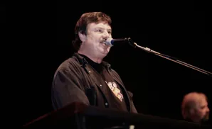 A man with a mustache sings into a microphone on stage, wearing a dark shirt and jacket.