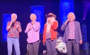 Four men singing on stage with microphones, wearing colorful shirts and jeans.