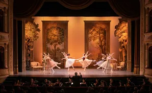 Ballet dancers perform on stage with an orchestra and audience in the foreground.
