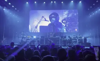 A musician performs on a stage in front of a large screen displaying their image, with a crowd watching.