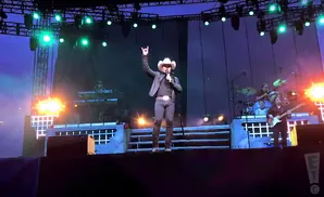 Country singer in a cowboy hat on stage at a concert, holding a microphone and making a rock-and-roll hand gesture.