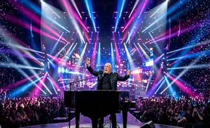 Billy Joel performs at a concert with vibrant lights and falling confetti as a large audience cheers him on.