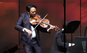 A man in a suit plays the violin on stage, with a pianist to his right.