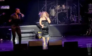 A male singer in a black shirt and pants sings into a microphone on stage next to a female singer in a black dress.