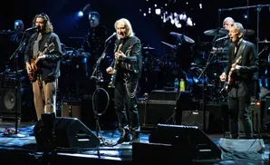 Musicians perform on stage with guitars, drums, and amplifiers, illuminated by blue stage lights.