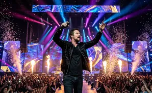 A male singer in a sparkly jacket performs on stage with confetti and lights, facing a cheering crowd.
