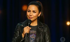 A woman with her hair in a high ponytail, wearing a black leather jacket and hoop earrings, holds a microphone and smiles.