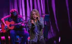 Robert Plant sings into a microphone on stage under purple lights, with a guitarist playing an acoustic guitar to his left.