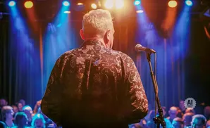 A man in a patterned shirt stands at a microphone on stage, facing away from the camera, with a blurry audience in the background.