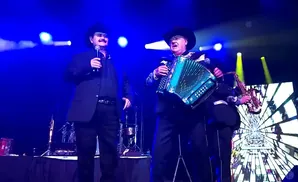 Two musicians perform on stage under blue lights. The one on the left sings into a microphone, while the one on the right plays an accordion and a saxop…
