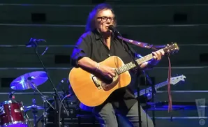 A person with glasses plays an acoustic guitar on stage, with drums and other instruments in the background.
