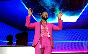John Legend in a bright pink suit with arms raised, singing on stage with a starry background.