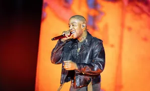 Rapper Kid Cudi performs on stage, wearing a black leather jacket and holding a red microphone.
