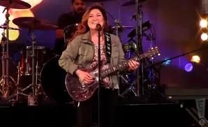 A woman with long brown hair sings and plays a purple electric guitar on stage.