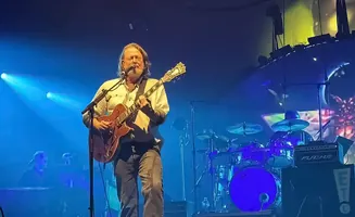 A man plays an electric guitar and sings on stage, with a drum set and keyboard player visible in the background.