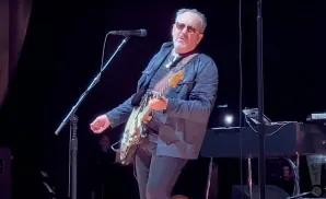 Elvis Costello plays guitar on stage, wearing sunglasses and a dark jacket.
