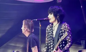 Joan Jett, in a star-print shirt, sings into a microphone while playing guitar on stage.