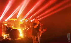 A singer on stage at a concert with red lights and a drum set behind him.