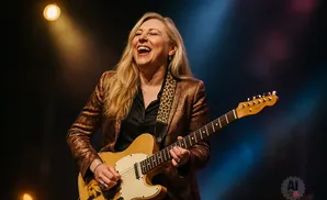 Woman laughing while playing a tan electric guitar on stage with colorful lights.