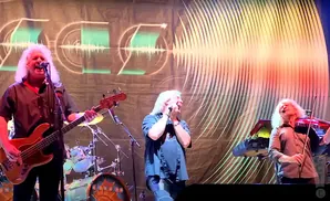 A band performs on stage, with a bassist in the foreground, a vocalist/keyboardist in the middle, and a violinist on the right.