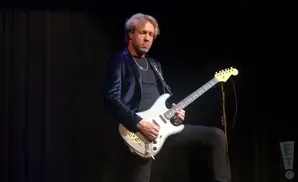 A man with blonde hair plays a silver electric guitar on a dark stage, wearing a dark velvet jacket.