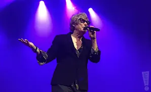 Man in black jacket and sunglasses sings into microphone on stage with purple lights.